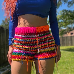 Könnte beinhalten: Mehrfarbige Häkelshorts mit einem rosa Bund. Die Shorts sind aus verschiedenen Garnfarben gefertigt, darunter Rot, Orange, Gelb, Grün, Blau und Lila.