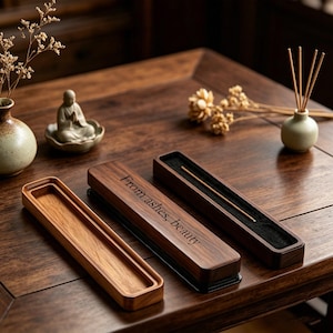 May include: Wooden incense holder with the engraved text "From ashes, beauty." The holder is rectangular and dark brown. The image also shows a small ceramic vase, a figurine, and dried flowers, all on a wooden table.