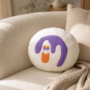 Puede incluir: Un coj&iacute;n decorativo redondo y blanco con un dise&ntilde;o de dibujos animados. El dise&ntilde;o presenta una forma morada con detalles naranjas, que se asemeja a una cara sonriente. El coj&iacute;n est&aacute; colocado en un sof&aacute; beige con una almohada a juego y una manta de punto.