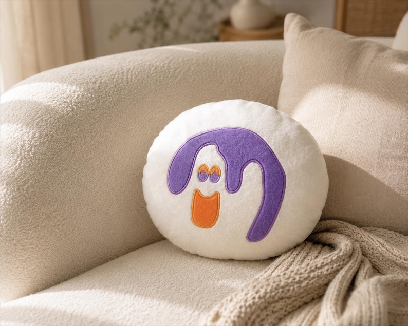 Puede incluir: Un coj&iacute;n decorativo redondo y blanco con un dise&ntilde;o de dibujos animados. El dise&ntilde;o presenta una forma morada con detalles naranjas, que se asemeja a una cara sonriente. El coj&iacute;n est&aacute; colocado en un sof&aacute; beige con una almohada a juego y una manta de punto.
