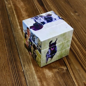 May include: A custom photo cube puzzle featuring images of a dog and people. The cube is white with images on each side, showcasing a playful dog in a field and a group of people. The puzzle sits on a wooden surface.