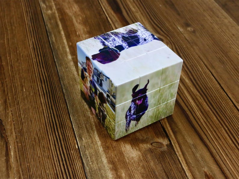 May include: A custom photo cube puzzle featuring images of a dog and people. The cube is white with images on each side, showcasing a playful dog in a field and a group of people. The puzzle sits on a wooden surface.