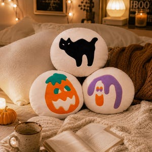 Puede incluir: Tres cojines redondos con tem&aacute;tica de Halloween. Un coj&iacute;n presenta una silueta de gato negro, otro una calabaza naranja y el tercero un fantasma morado. Los cojines son blancos con aplicaciones coloridas, descansando sobre una manta acogedora.
