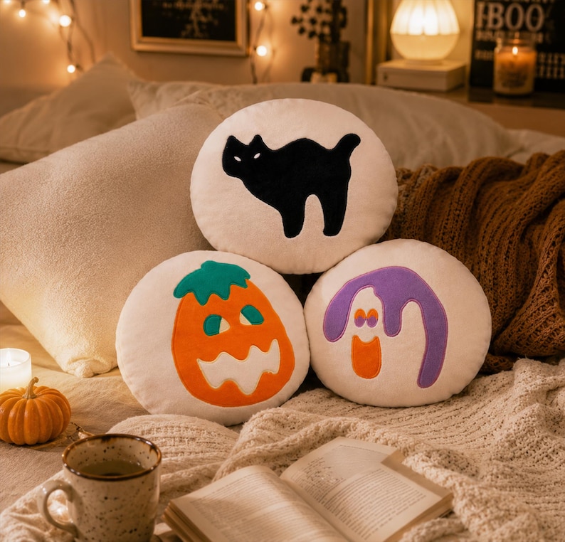 Puede incluir: Tres cojines redondos con tem&aacute;tica de Halloween. Un coj&iacute;n presenta una silueta de gato negro, otro una calabaza naranja y el tercero un fantasma morado. Los cojines son blancos con aplicaciones coloridas, descansando sobre una manta acogedora.