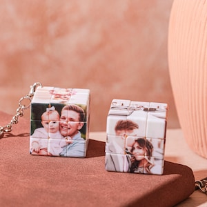 Può includere: Due portachiavi a cubo di Rubik bianchi con foto personalizzate. Un cubo mostra un uomo e un bambino, l'altro una coppia. I cubi sono su una superficie marrone con uno sfondo rosa. Una catena d'argento è attaccata a un cubo.