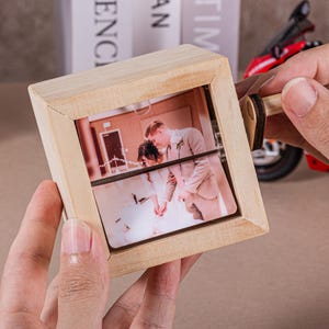 May include: A wooden photo cube with a rotating mechanism, displaying a wedding photo. The cube is made of light-coloured wood and has a square frame. The photo shows a bride and groom. A small red toy motorcycle is in the background.