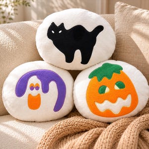 Puede incluir: Tres cojines redondos con tem&aacute;tica de Halloween. Uno presenta una silueta de gato negro, otro un fantasma morado con detalles en naranja y el tercero una calabaza naranja con un tallo verde. Los cojines son blancos y est&aacute;n sobre un sof&aacute; beige.