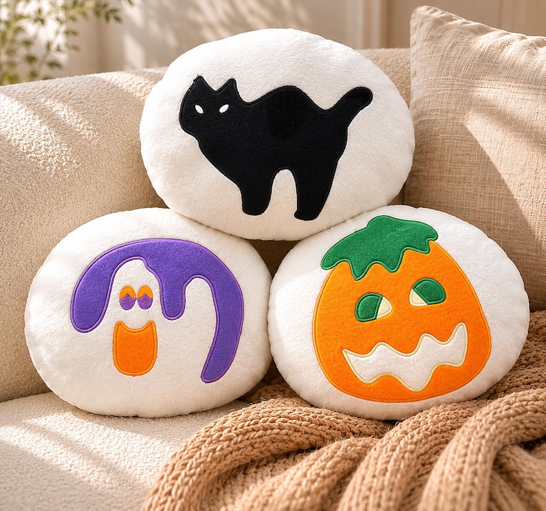 Puede incluir: Tres cojines redondos con tem&aacute;tica de Halloween. Uno presenta una silueta de gato negro, otro un fantasma morado con detalles en naranja y el tercero una calabaza naranja con un tallo verde. Los cojines son blancos y est&aacute;n sobre un sof&aacute; beige.