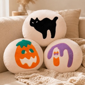 May include: Three round Halloween-themed throw pillows. One features a black cat silhouette, another an orange jack-o'-lantern with a green stem, and the third a purple ghost with an orange mouth. The pillows are on a beige couch.