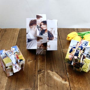 May include: Three photo cubes, each displaying multiple images, sit on a wooden surface. The central cube is a larger, multi-faceted structure. The other two cubes are smaller and show various photographs. Yellow tulips and green leaves are in the background.