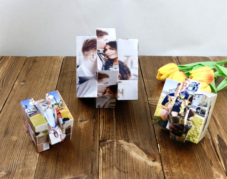 May include: Three photo cubes, each displaying multiple images, sit on a wooden surface. The central cube is a larger, multi-faceted structure. The other two cubes are smaller and show various photographs. Yellow tulips and green leaves are in the background.