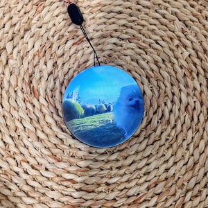 Può includere: Un ciondolo rotondo blu con un cane bianco e soffice su uno sfondo di campo verde e castello. Il ciondolo è appeso a un cordino nero e a un cordino intrecciato rosa e beige.