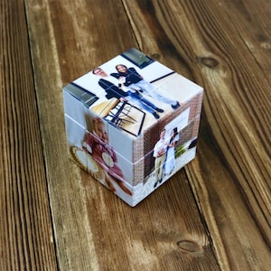 May include: A personalized photo cube puzzle with images of people and brick wall patterns. The cube sits on a wooden surface, showcasing the custom printed photos on each side. A unique gift idea.
