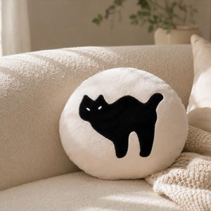 Puede incluir: Un coj&iacute;n decorativo redondo de color crema con una silueta de gato negro. El gato est&aacute; arqueado con la cola levantada, y el coj&iacute;n est&aacute; sobre un sof&aacute; beige. Tambi&eacute;n se ve una manta beige y otros cojines.