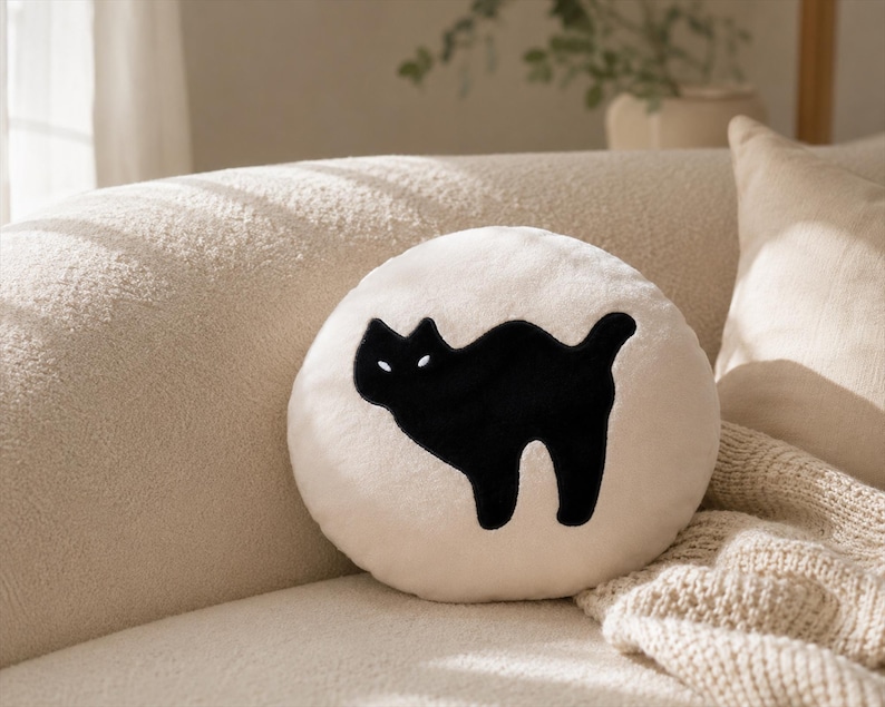 Puede incluir: Un coj&iacute;n decorativo redondo de color crema con una silueta de gato negro. El gato est&aacute; arqueado con la cola levantada, y el coj&iacute;n est&aacute; sobre un sof&aacute; beige. Tambi&eacute;n se ve una manta beige y otros cojines.