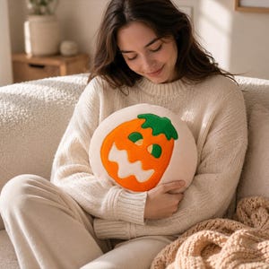 Puede incluir: Un coj&iacute;n redondo de color crema con un dise&ntilde;o de cara de calabaza naranja. La calabaza tiene un tallo verde y una cara sonriente. El coj&iacute;n lo sostiene una persona que lleva un su&eacute;ter color crema.