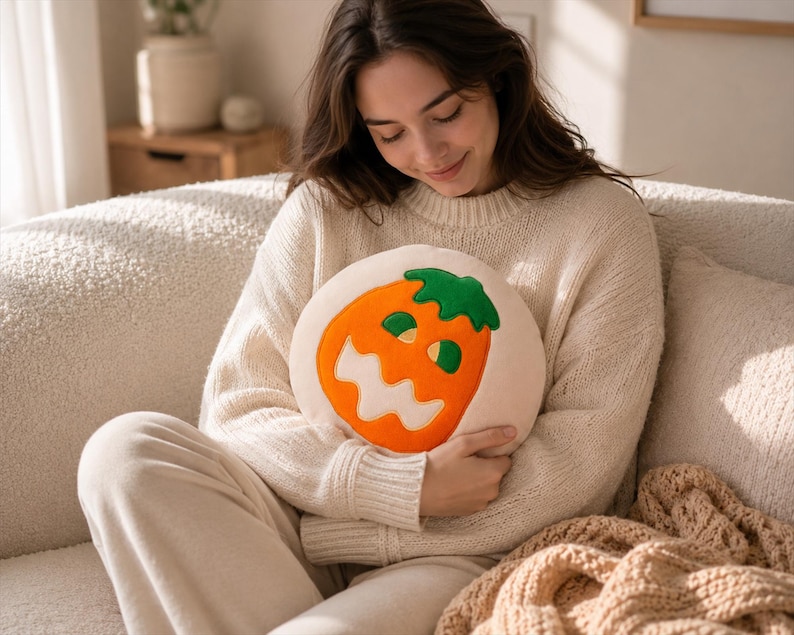 Puede incluir: Un coj&iacute;n redondo de color crema con un dise&ntilde;o de cara de calabaza naranja. La calabaza tiene un tallo verde y una cara sonriente. El coj&iacute;n lo sostiene una persona que lleva un su&eacute;ter color crema.