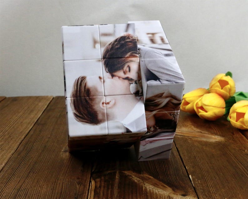 May include: A personalized photo cube featuring images of a couple kissing. The cube is made of white material and displays the photos on its sides. Yellow tulips are in the background.