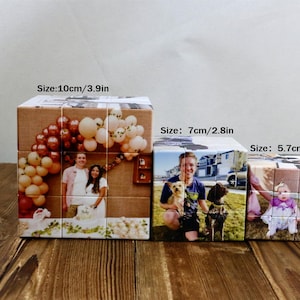 May include: Three personalized photo cubes of varying sizes (10cm/3.9in, 7cm/2.8in, and 5.7cm/2.2in) sit on a wooden surface. Each cube displays a different photograph, including a wedding photo, a person with dogs, and a baby.