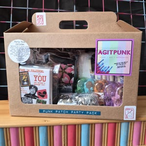Puede incluir: Una caja de cartón marrón etiquetada como Punk Patch Party Pack que contiene varios suministros para manualidades, incluyendo un paquete de tarjetas con el texto Punk is Political, una tela floral rosa, tachuelas metálicas y clips de plástico.
