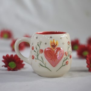 Könnte beinhalten: Eine weiße Keramiktasse mit rotem Innenraum und Herzdesign. Das Herz ist rot mit einer Flamme oben, umgeben von floralen und sternförmigen Details. Die Tasse hat einen gebogenen Griff und ist mit goldenen Akzenten verziert.