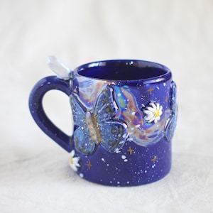 Könnte beinhalten: Eine dunkelblaue Keramiktasse mit Henkel und Kristallakzent. Die Tasse zeigt geprägte Schmetterlinge, Gänseblümchen und Sterne. Das Design umfasst schillernde und weiße Akzente und schafft ein skurriles, himmlisches Thema.