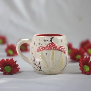 Könnte beinhalten: Cremefarbene Keramiktasse mit rotem Innenraum, verziert mit einem Pilzdesign. Die Pilze haben rote Kappen mit weißen Punkten. Die Tasse ist mit Sternen und einem Mond verziert. Ideal für Tee oder Kaffee.