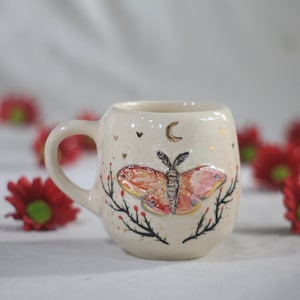 Könnte beinhalten: Cremefarbene Keramiktasse mit Henkel, verziert mit einem bunten Motten-Design. Die Motte hat orange, rosa und braune Flügel. Die Tasse hat auch einen goldenen Halbmond, Herzen und kleine goldene Punkte.