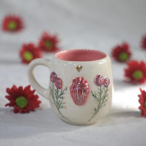 Könnte beinhalten: Cremefarbene Keramik-Tasse mit rosa Innenraum und Henkel. Die Tasse ist mit einem erhabenen, strukturierten anatomischen Herzen und floralen Designs in Rot- und Lilatönen verziert. Ein kleines goldenes Herz befindet sich in der Nähe des Randes.