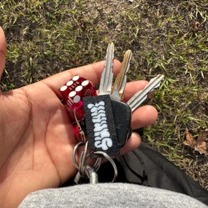 May include: A red dice keychain with the numbers 2, 3, 7, and 6 on the sides. The keychain is attached to a black key holder with the word "SHATTER" written in white letters. The keychain is attached to a set of keys.
