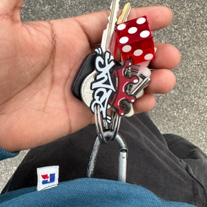 May include: A hand holding a set of keys with a red dice charm and a silver carabiner. The keys have a black and white charm with the word "King" written in graffiti style.