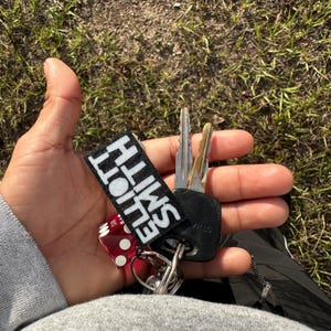 May include: A black key chain with a red dice charm and a black patch with white text that reads "SMITH  HOLT".