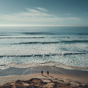 May include: Two people walk along a sandy beach towards the ocean. The water is a light blue and the sky is a clear blue.