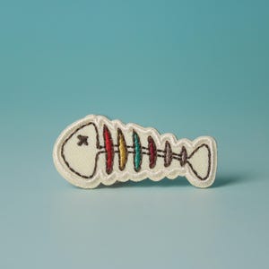 Könnte beinhalten: Ein weißer Filz-Fisch-Patch mit einem Regenbogen-Skelett-Design. Der Fisch hat ein schwarzes Auge und ein kleines schwarzes X auf dem Kopf.