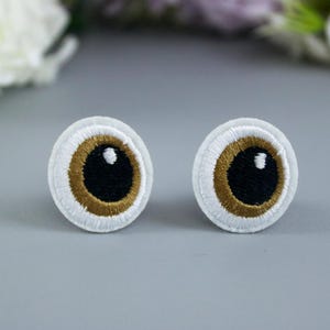 May include: Two embroidered felt eyes with brown irises and white pupils. The eyes are on a white background.