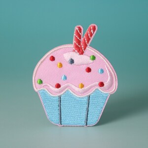 Puede incluir: Un parche bordado de cupcake rosa y azul con velas rojas y blancas y chispas de colores.