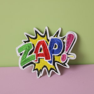 Może przedstawiać: Naszywka do prasowania w kolorze żółtym, czerwonym, niebieskim i zielonym z haftowanym napisem "ZAP!" w stylu komiksowym.