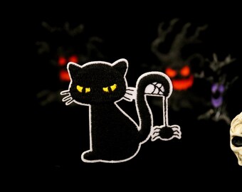 Op de afbeelding: Zwarte kattenpatch met gele ogen en witte omtrek, die een spin in zijn staart vasthoudt. De patch staat tegen een donkere achtergrond met Halloween-decoraties, waaronder een schedel.