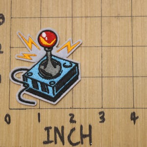 May include: An embroidered patch of a blue and grey joystick with a red button and yellow lightning bolts. The patch is approximately 5 cm wide.