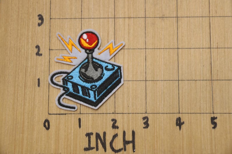 May include: An embroidered patch of a blue and grey joystick with a red button and yellow lightning bolts. The patch is approximately 5 cm wide.