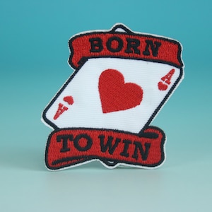 Puede incluir: Parche bordado rojo y blanco con un diseño de carta de juego. La carta es un as de corazones con el texto "BORN TO WIN" en una pancarta roja.