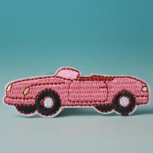 May include: Pink embroidered iron-on patch of a convertible car. The car has black wheels and white headlights.