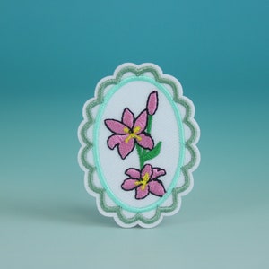 Puede incluir: Un parche ovalado blanco bordado con un borde verde festoneado. El parche presenta una flor rosa con un centro amarillo y hojas verdes.