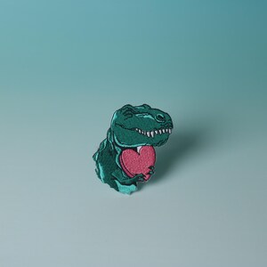 Pode incluir: Um dinossauro verde segurando um cora&ccedil;&atilde;o rosa. O dinossauro est&aacute; sorrindo e tem uma express&atilde;o amig&aacute;vel. O cora&ccedil;&atilde;o &eacute; uma forma simples sem detalhes.