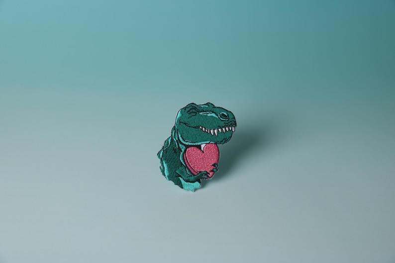 Pode incluir: Um dinossauro verde segurando um cora&ccedil;&atilde;o rosa. O dinossauro est&aacute; sorrindo e tem uma express&atilde;o amig&aacute;vel. O cora&ccedil;&atilde;o &eacute; uma forma simples sem detalhes.