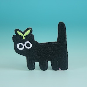 May include: Black felt cat with white eyes and a green sprout on its head. The cat is standing on a light blue background.
