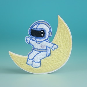 May include: A white astronaut with a blue helmet sits on a yellow crescent moon. The astronaut has a black visor and is wearing a white spacesuit with blue accents.