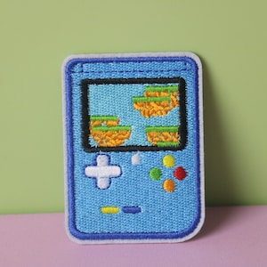 May include: A blue embroidered patch of a handheld video game console with a pixelated game on the screen. The console has a white plus button and four colored buttons.