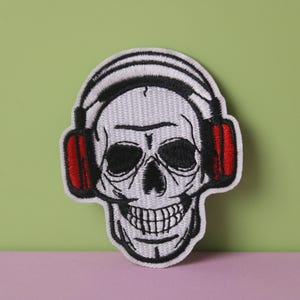 Puede incluir: Cráneo blanco con ojos negros y una sonrisa de dientes, con auriculares rojos. El cráneo está bordado en un parche de tela blanca.