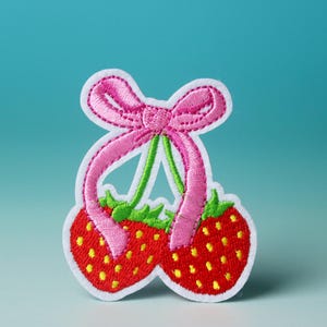 Puede incluir: Parche bordado con dos fresas rojas con hojas verdes y un lazo rosa.